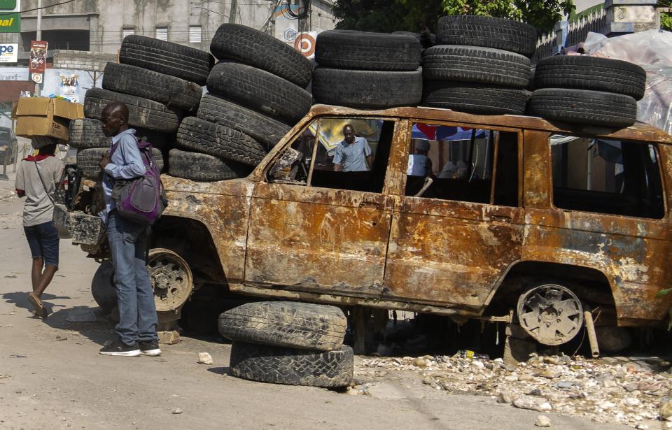 Gente pasa junto a neumáticos y un vehículo quemado utilizado para cortar una calle en la zona de Canapé Vert, Puerto Príncipe, Haití, el lunes 22 de abril de 2024. Hace mucho que el sistema de salud de Haití era frágil, pero ahora se acerca al colapso total después de que las pandillas lanzaran ataques coordinados el 29 de febrero contra infraestructura crítica en la capital y otros lugares.. (AP Foto/Ramón Espinosa)