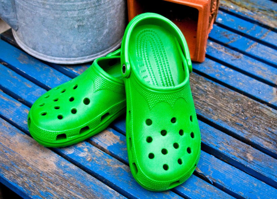 A pair of green Crocs is placed on a blue wooden surface next to a metal bucket and a piece of clay pottery