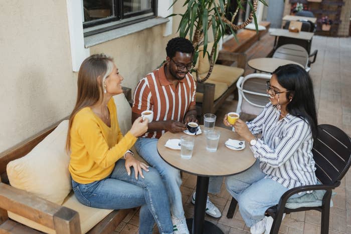 Three people sit at an outdoor café table, enjoying coffee and conversation. The group is casually dressed