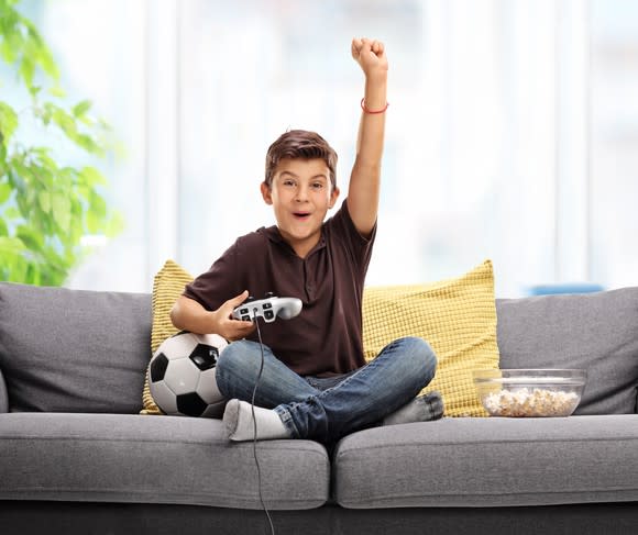A boy sitting on a couch next to a soccer ball and a bowl of popcorn, and holding a video game controller, smiling with his arm reaching up into the air.