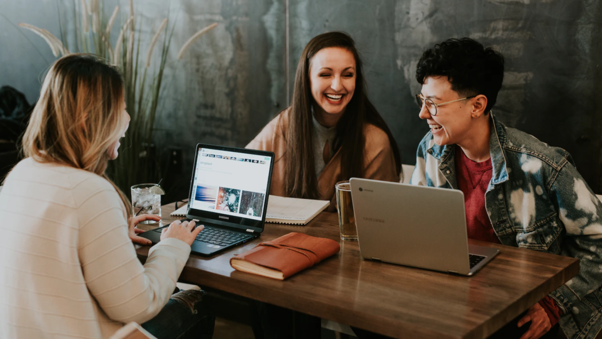  Three women with laptops sitting around a table laughing. 