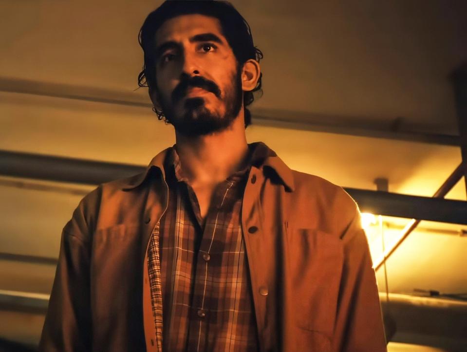 in a scene, Dev Patel in a dimly lit indoor setting, wearing a plaid shirt under a jacket