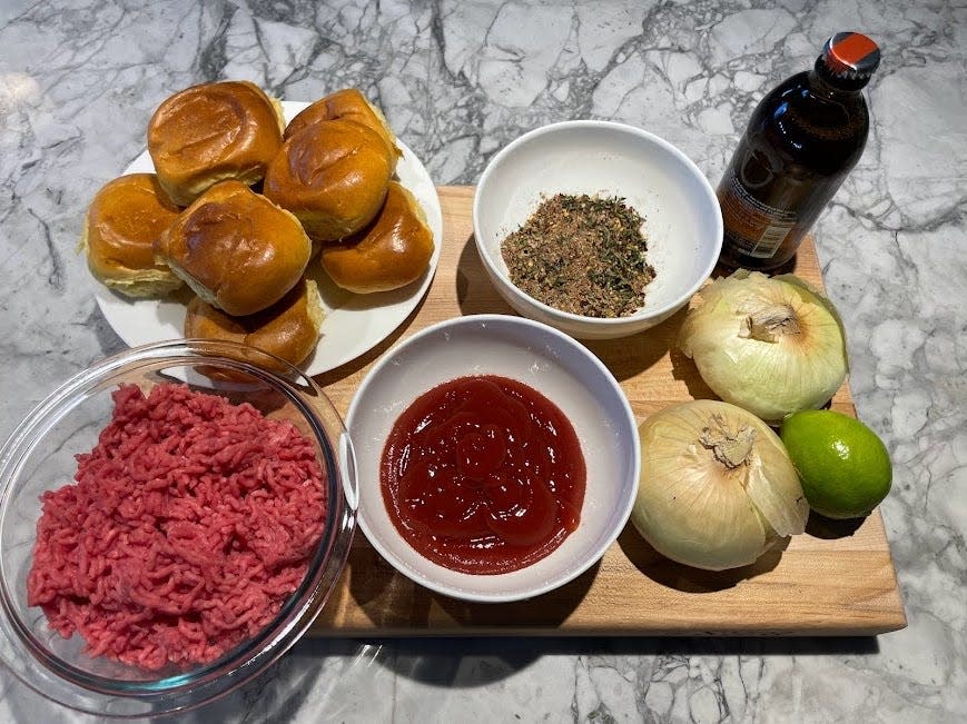 A wooden cutting board with hamburger buns, ground beef, ketchup, spice mixture, a bottle of beer, two onions, and a lime