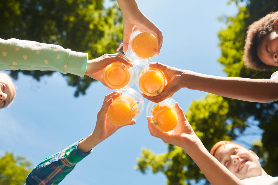 Low angle close up of kids holding glasses with orange juice against blue sky outdoors, copy space