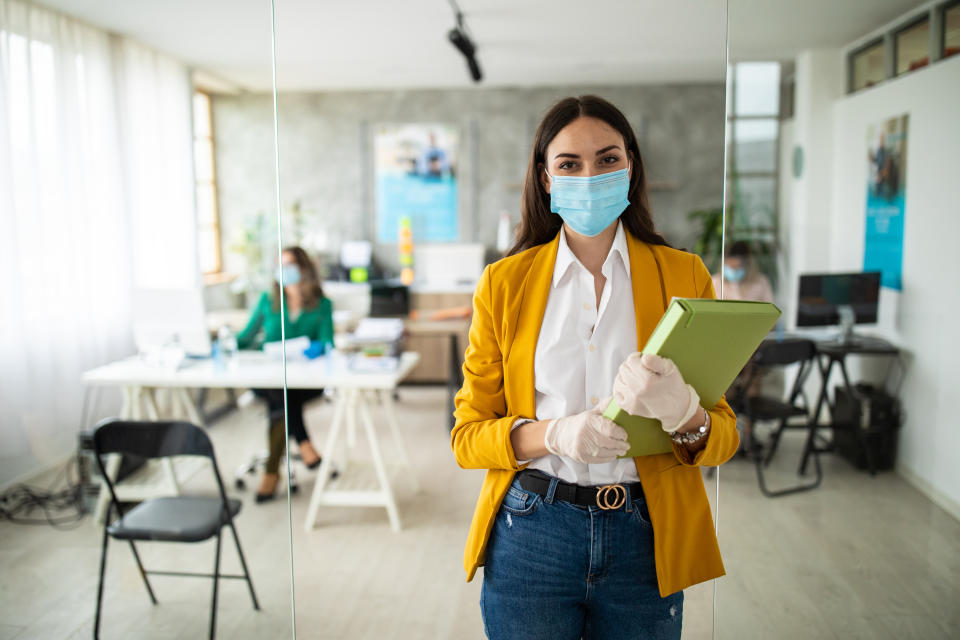 Portrait of female bank manager with protective face mask and surgical gloves in office, standing, looking at camera and smiling during covid-19 pandemic