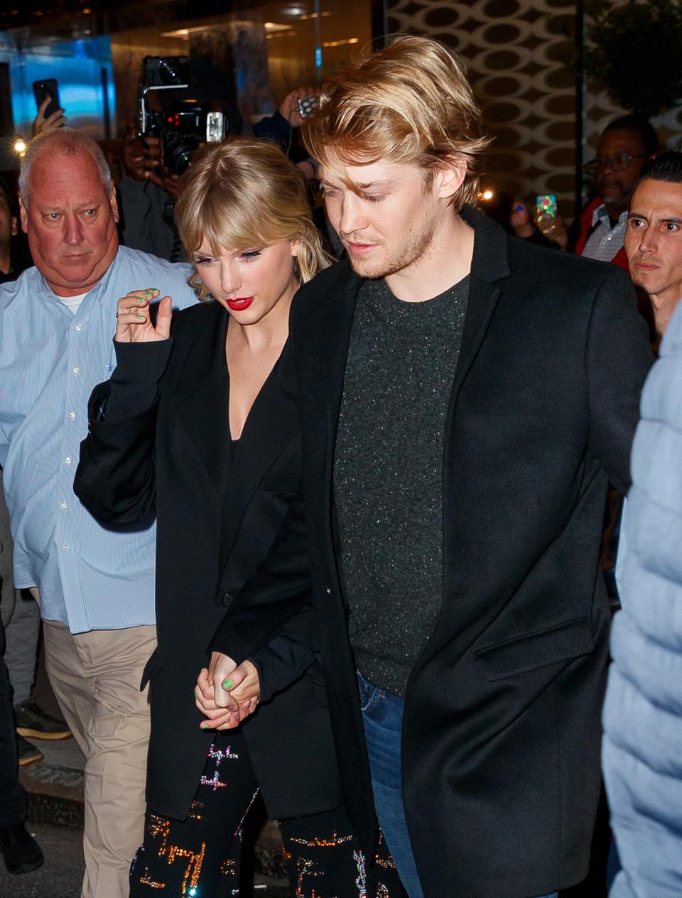 Taylor Swift and Joe Alwyn walking together, she in a sequined outfit, he in a dark coat. Photographers in the background