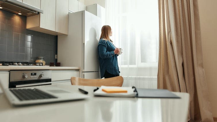 Woman stands at her kitchen window with a mug of tea or coffee.