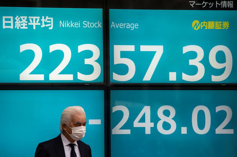 A man wearing a protective face mask stands in front of a screen showing the Nikkei index in Tokyo