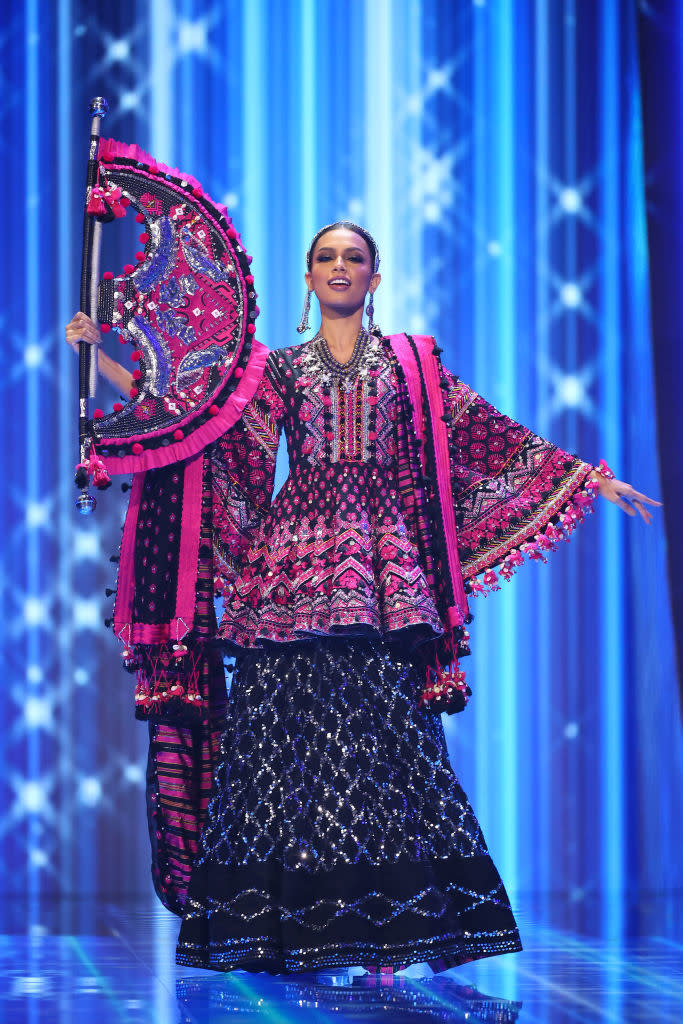 She's wearing a colorful bejeweled, tiered gown with flared sleeves and ornate matching fan