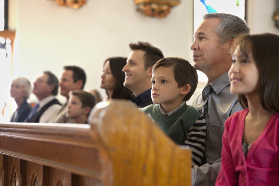 A group of people, including children and adults, sits attentively in what appears to be a church pew, looking towards the front