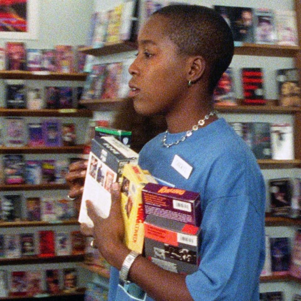 A person in a store carries multiple VHS tapes, wearing a short-sleeve top and a necklace in 