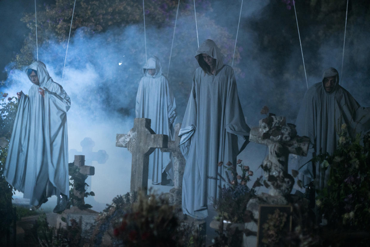 Four people in gray robes hang, suspended from ropes, above a cemetery.