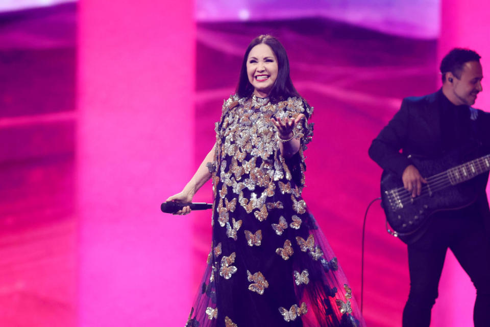 Ana on stage, smiling and holding a microphone, wearing a dress adorned with butterfly designs, with a band member playing guitar in the background