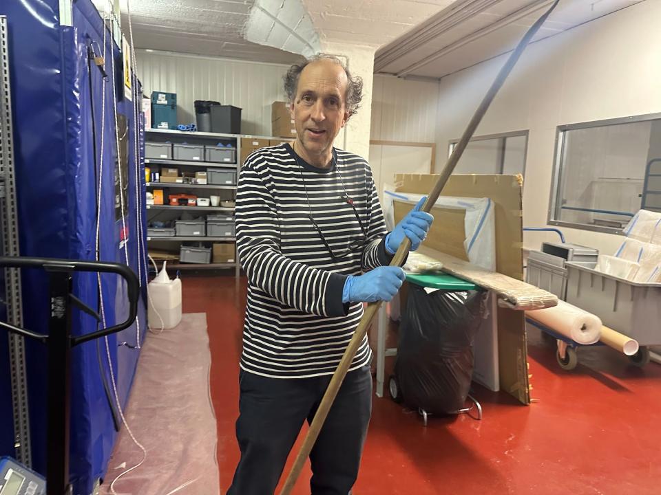 A man wearing gloves holds a long wooden bow in a basement room full of storage bins and packing supplies.