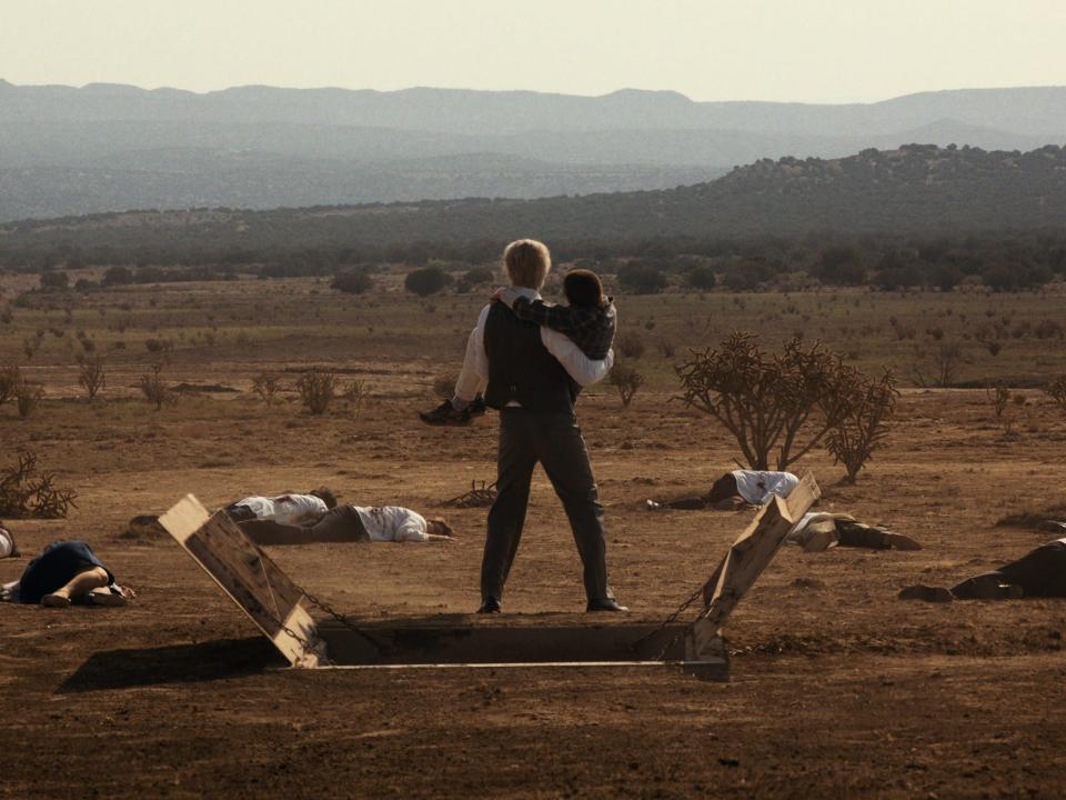 a man carrying a teenage girl, standing in the sand in a desert with bodies wearing white coats strewn around him in stranger things