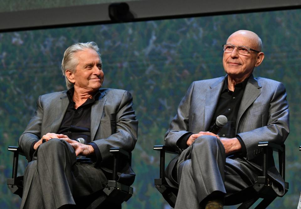 michael douglas and alan arkin, wearing gray suits and black shirts, sitting on chairs on a stage