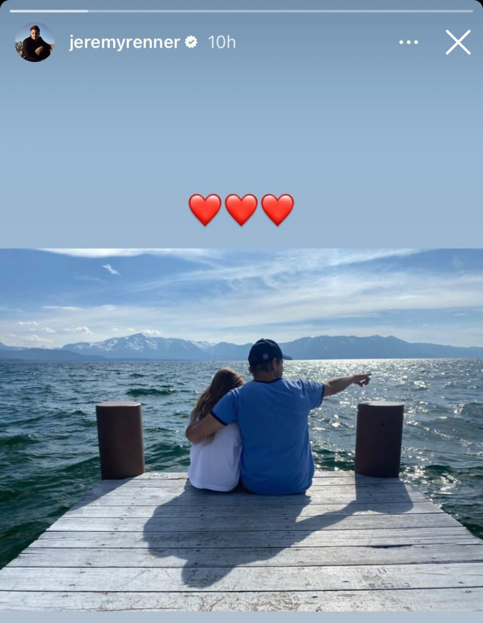 Renner sits at the end of a dock overlooking a lake with mountains in the distance. He points to the right with his right hand, while his left hand is wrapped around his daughter. (Jeremy Renner / Instagram)