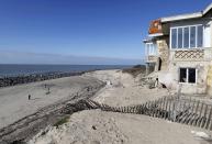 Varias personas caminan a lo largo de la playa frente a una casa dañada donde las dunas de arena no han podido parar la erosión.