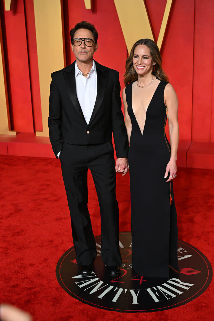 Robert Downey Jr. and Susan Downey standing together; he's in a classic suit, she's in a black evening gown with a plunge neckline