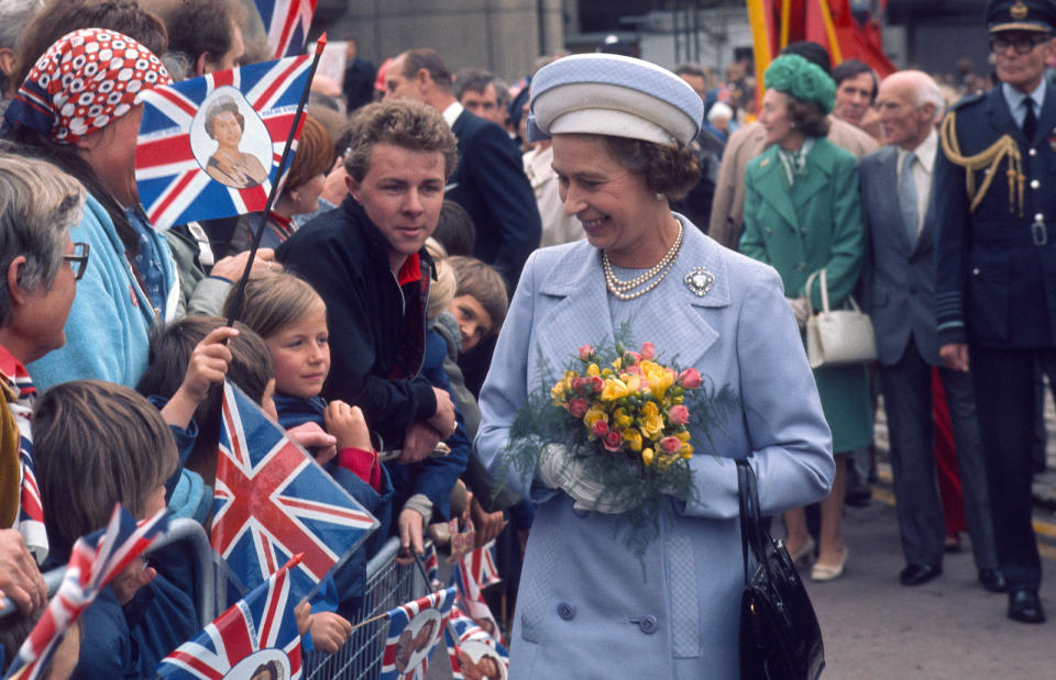 Queen Elizabeth ll greets the public during a Silver Jubilee walkabout wearing a pale blue double-breasted long coat, pearls and hat, carrying a bunch of roses.