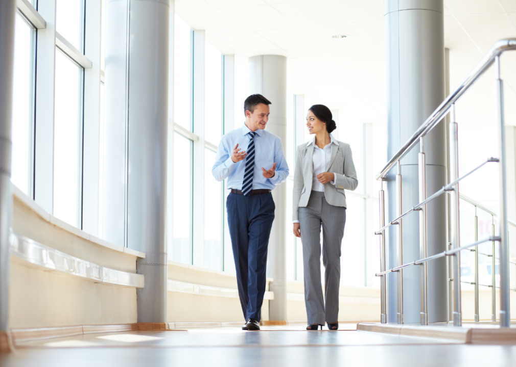 Two businesspeople walk down a brightly lit hallway while talking. 