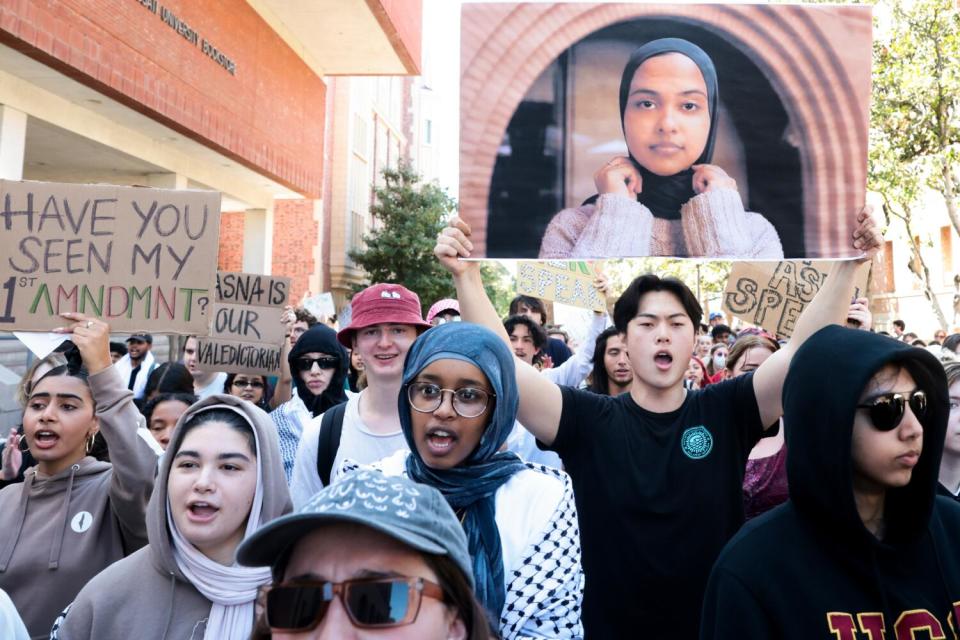 A crowd marching, one holding a large photo of Asna Tabassum, another a sign reading "Have you seen my 1st Amndmnt?"