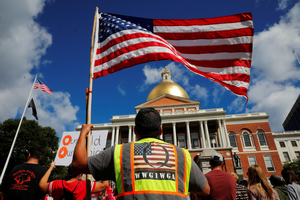 A man wearing a QAnon vest holding an American flag demonstrates outside the State House in Boston, Mass.