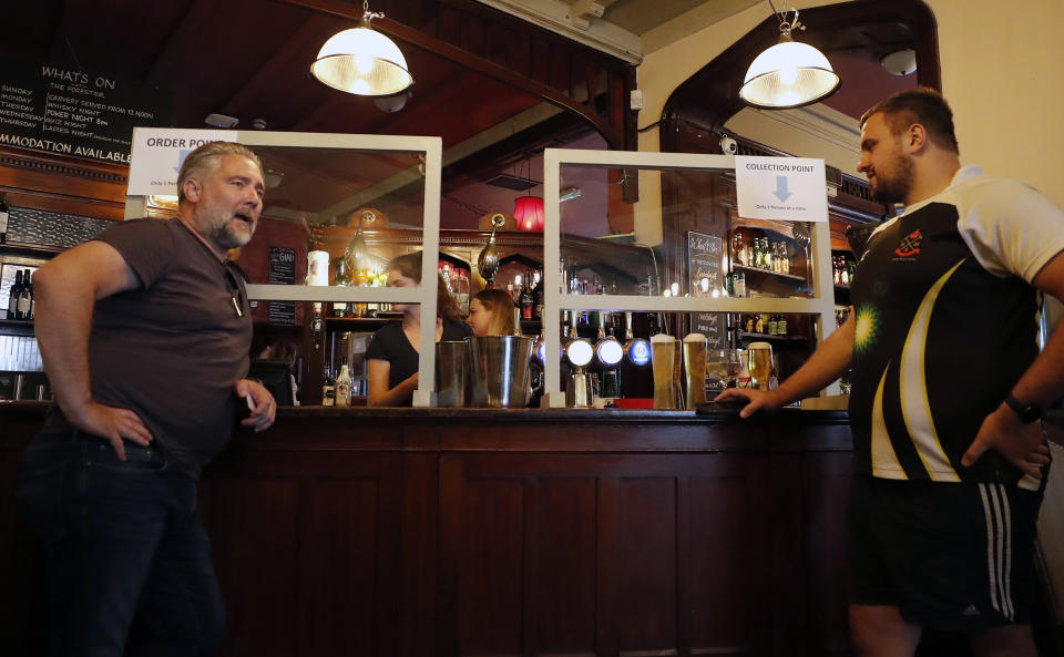 Men order beer behind a safety screen at the Forester pub in London, Saturday, July 4, 2020. England is embarking on perhaps its biggest lockdown easing yet as pubs and restaurants have the right to reopen for the first time in more than three months. In addition to the reopening of much of the hospitality sector, couples can tie the knot once again, while many of those who have had enough of their lockdown hair can finally get a trim. (AP Photo/Frank Augstein)