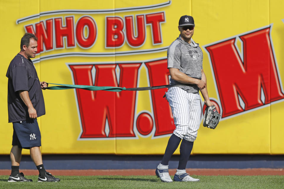 New York Yankees' Giancarlo Stanton, right, who has been out the much of the season with various injuries, takes a break as he works with a trainer and a resistance band in the outfield before a baseball game between the Yankees and the Texas Rangers, Wednesday, Sept. 4, 2019, in New York. (AP Photo/Kathy Willens)