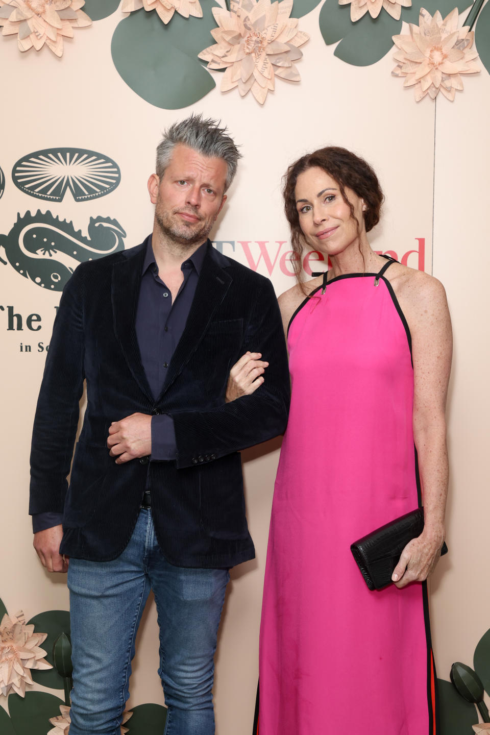 Addison O'Dea and Minnie Driver standing together. The man wears a dark blazer over a shirt and blue jeans. The woman wears a sleeveless pink dress, holding a black clutch