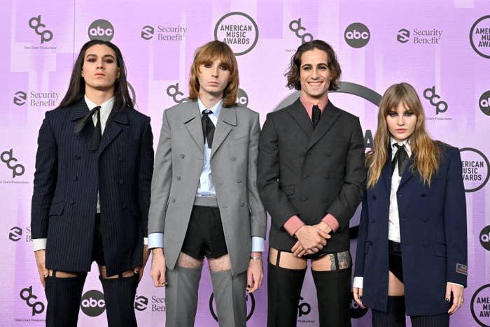 The members of Måneskin posing for a group photo at the American Music Awards. They're all wearing suit jackets and ties along with dress shirts and very short shorts and stockings held up by garter belts