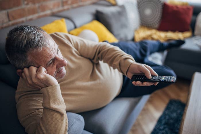 A man lies on a couch holding a TV remote, appearing relaxed and comfortable in a casual setting