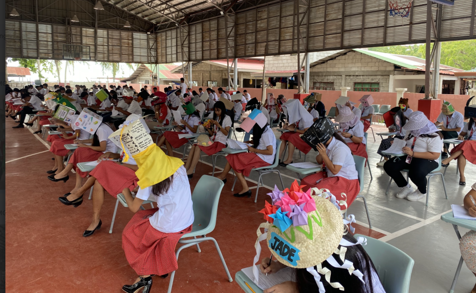 Students sitting with books covering their faces, some wearing decorated hats, in an open-air covered area