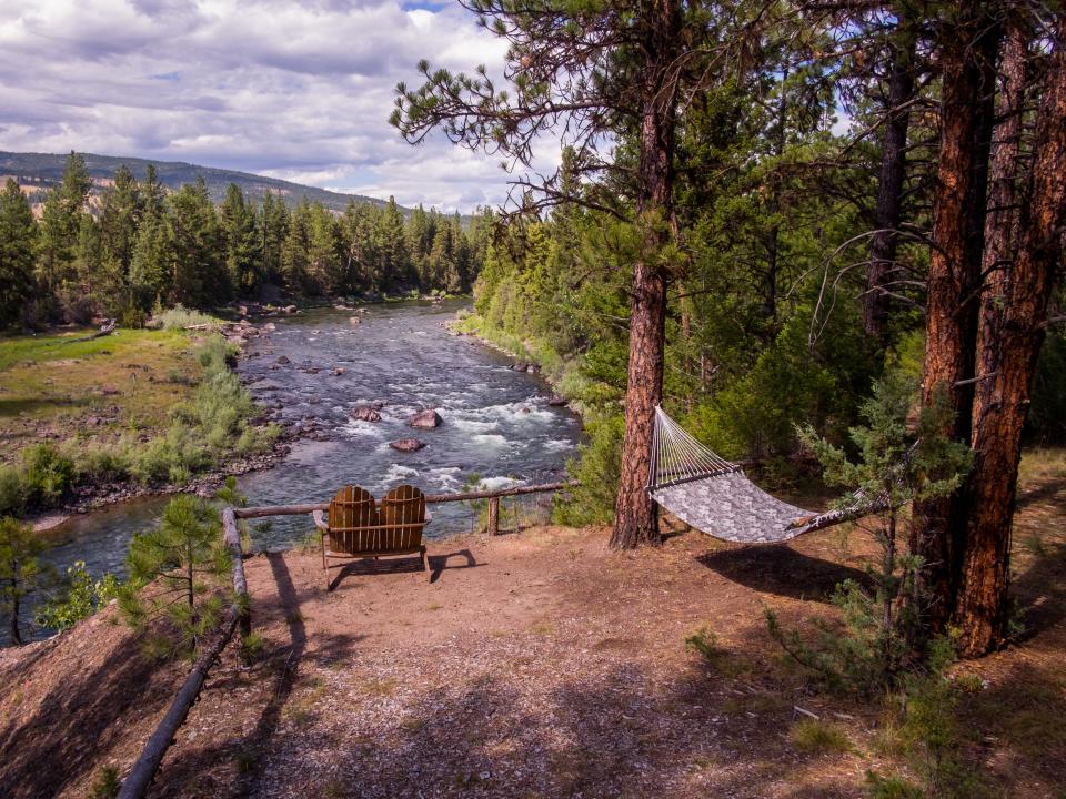 A river surrounded by trees and mountains, with two chairs and a hammock overlooking it.