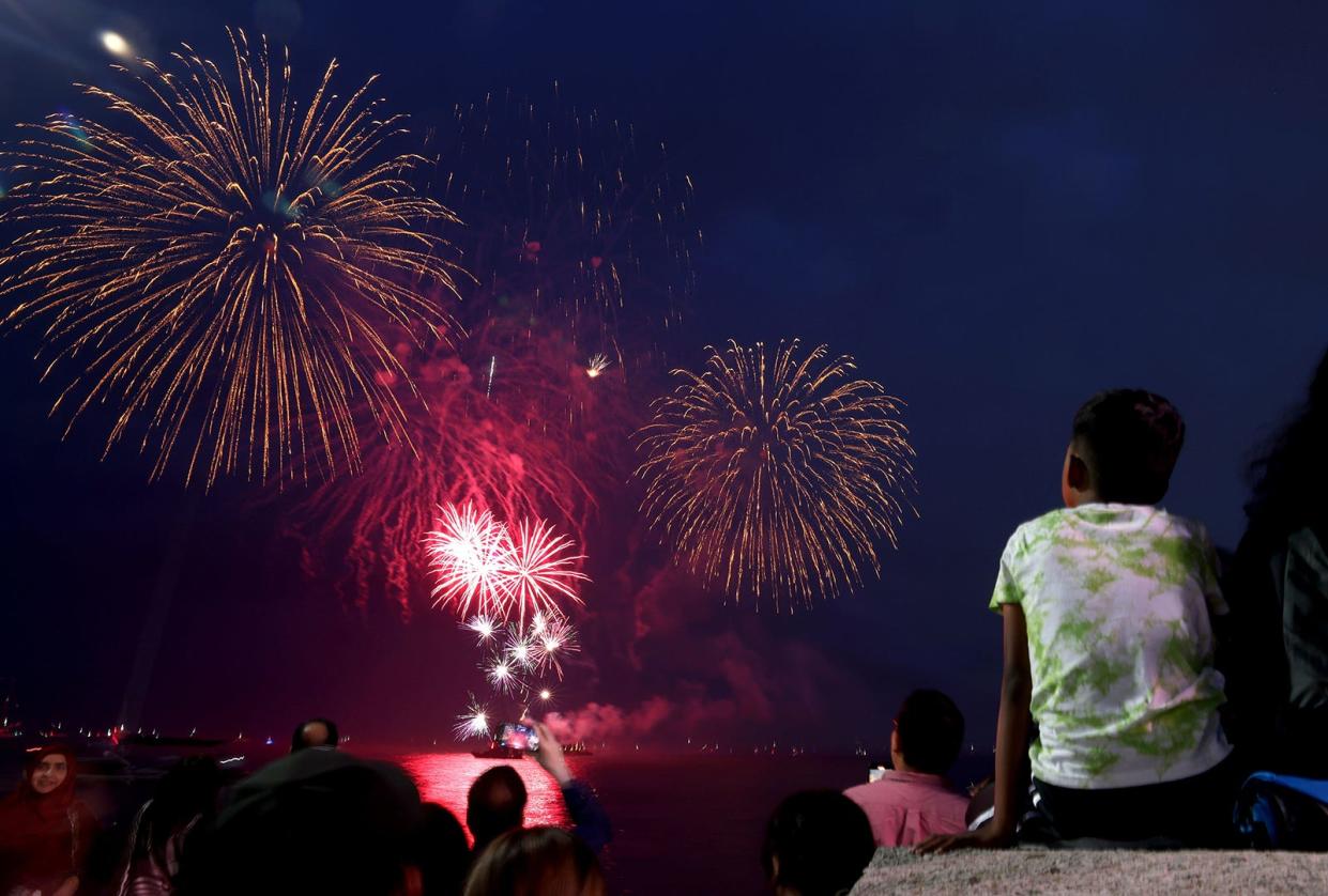 A group of people gathered together near a pier while a barge sets off fireworks in the distance.