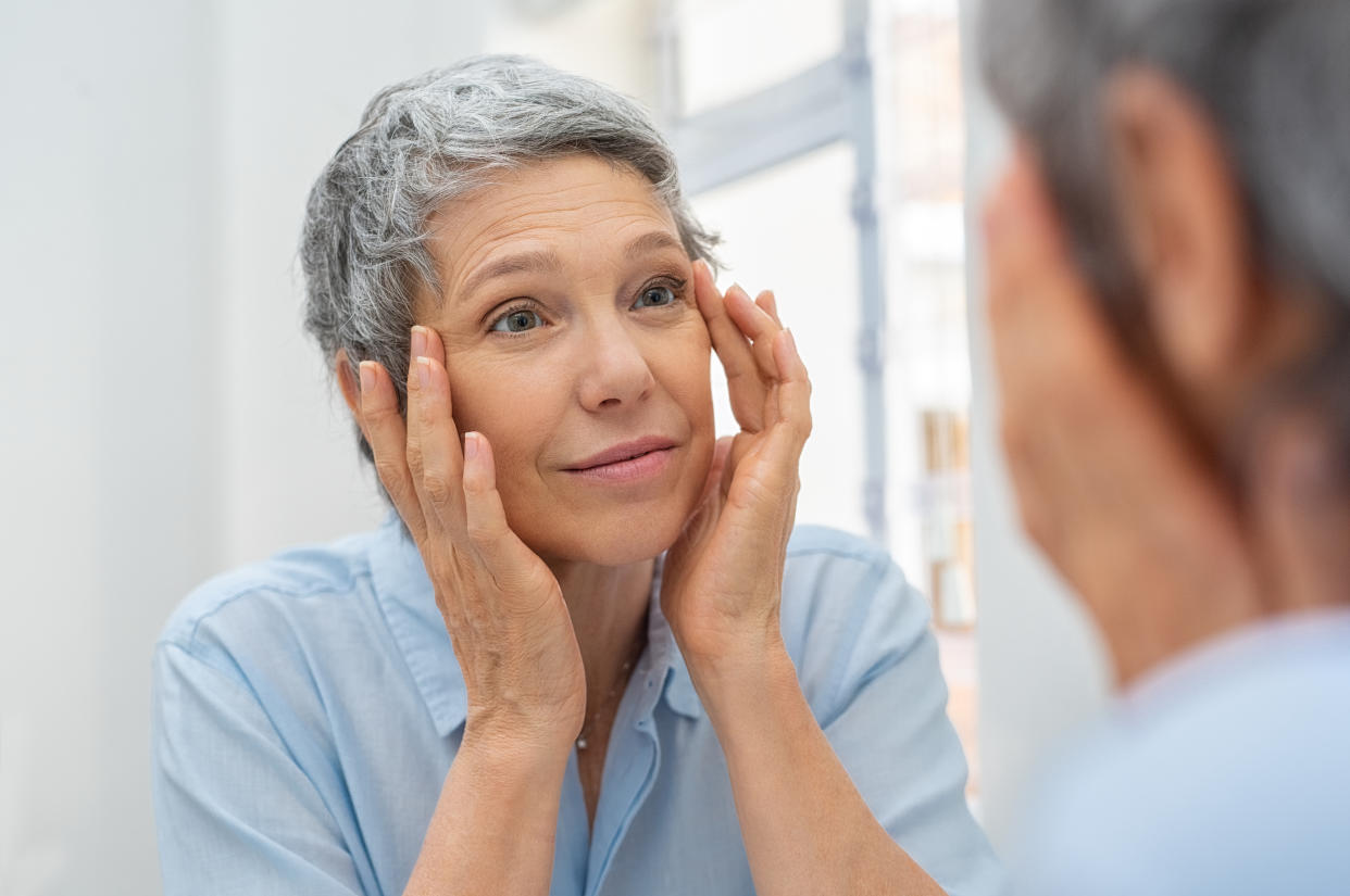 Beautiful senior woman checking her face skin and looking for blemishes. Portrait of mature woman massaging her face while checking wrinkled eyes in the mirror. Wrinkled lady with grey hair checking wrinkles around eyes, aging process.