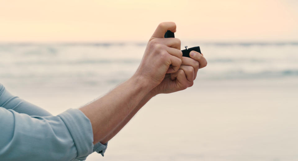 An unrecognizable man on bended knee, showing an open box with an engagement ring in it at the beach. Closeup of a man making a wedding proposal