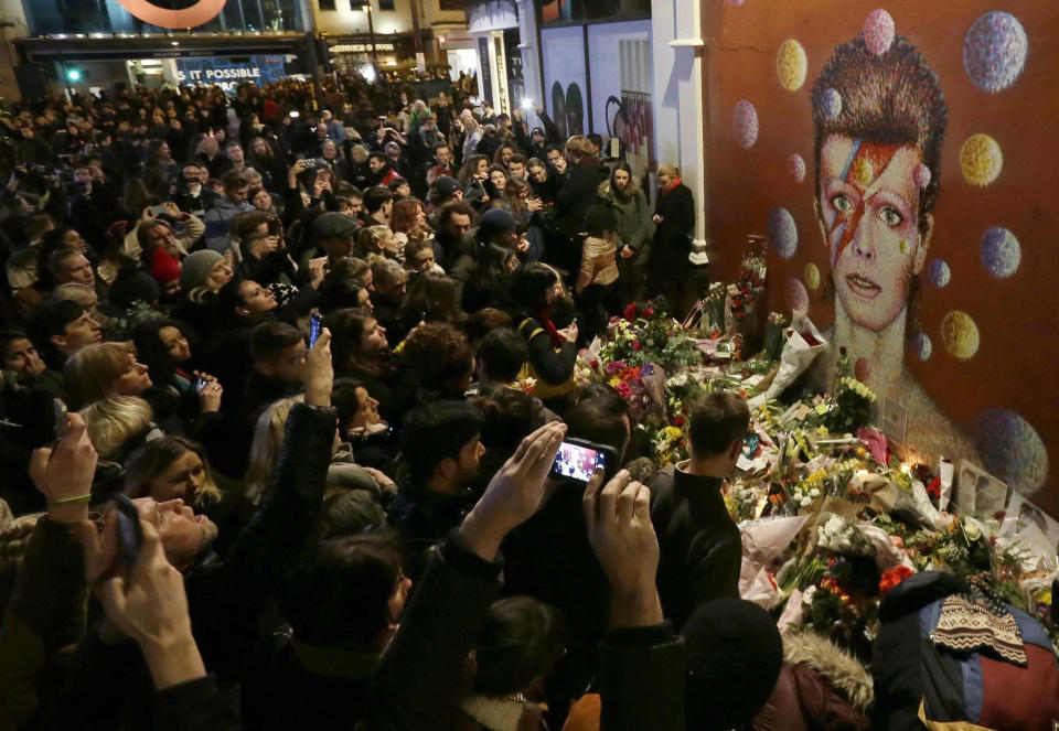 FILE - In this Monday, Jan. 11, 2016 file photo people gather next to tributes placed near a mural of British musician David Bowie by artist Jimmy C, in Brixton, south London. Bowie, the other-worldly musician who broke pop and rock boundaries with his creative musicianship, nonconformity, striking visuals and a genre-spanning persona he christened Ziggy Stardust, died Jan. 10, 2016. (AP Photo/Tim Ireland, File)