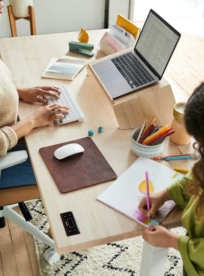 woman works on laptop while a child is drawing on the same table nearby