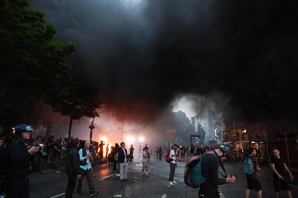 A black cloud moves through the air as demonstrators walk in the street while a fire burns in town.&nbsp;