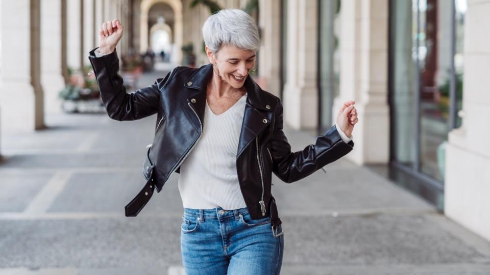 A woman with grey hear wearing a black leather jacket and jeans dancing while she walks