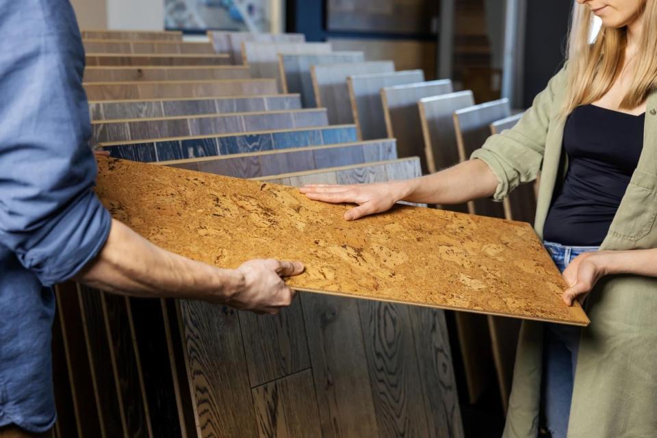 A man shows a sheet of cork flooring to a woman to feels it with her hand. 
