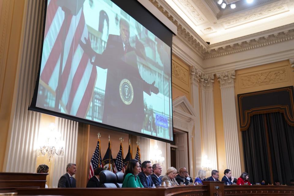 A video of President Trump addressing a crowd outside the White House plays on a large screen at the front of a hearing room.
