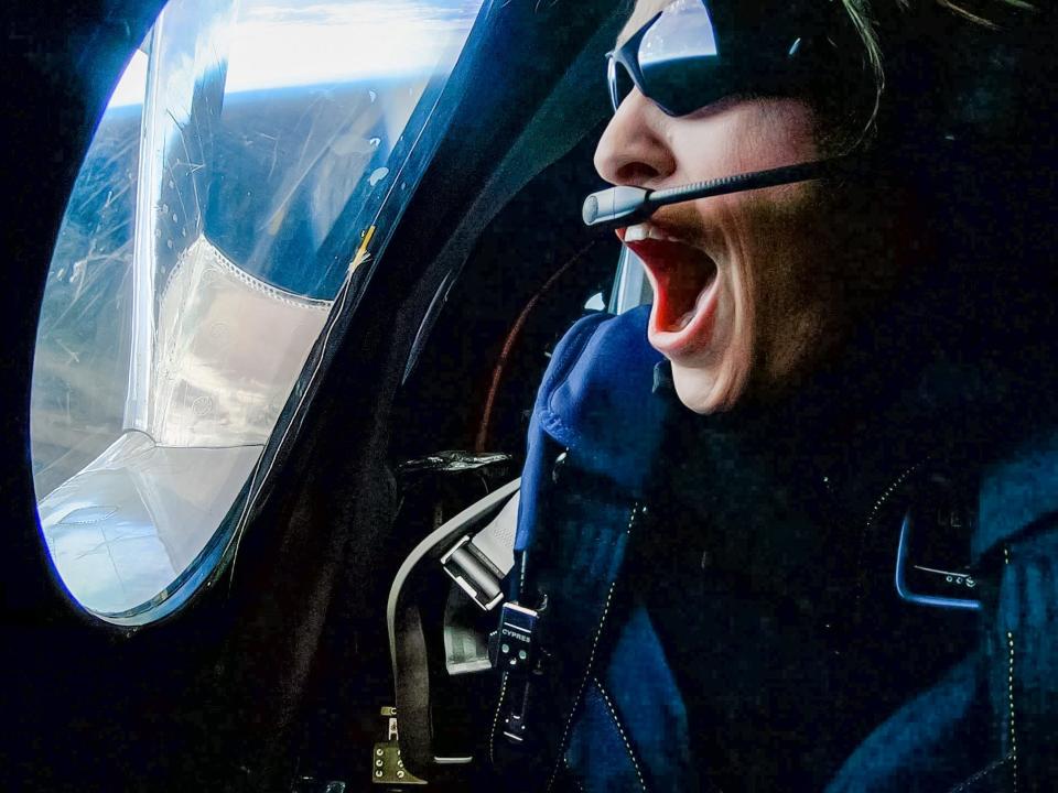 A woman with sunglasses and a headset opening her mouth and looking out the window of a space plane.