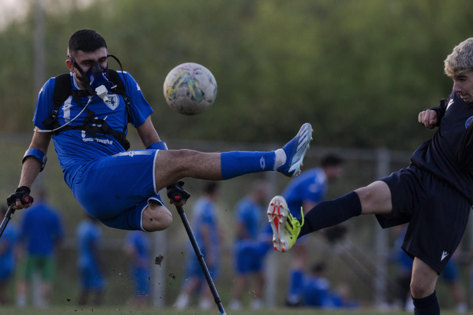 Wearing a device that measures his energy consumption, the Israel Amputee Football Team player, Ben Maman, left, fights for the ball with a young soccer player from a local team during a practice session in Ramat Gan, Thursday, April 11, 2024. Amputee football stands out as a disability sport because the athletes aren't in wheelchairs. It is played with six outfield players who have lower extremity amputations and play with crutches and without prosthetics. (AP Photo/Leo Correa)