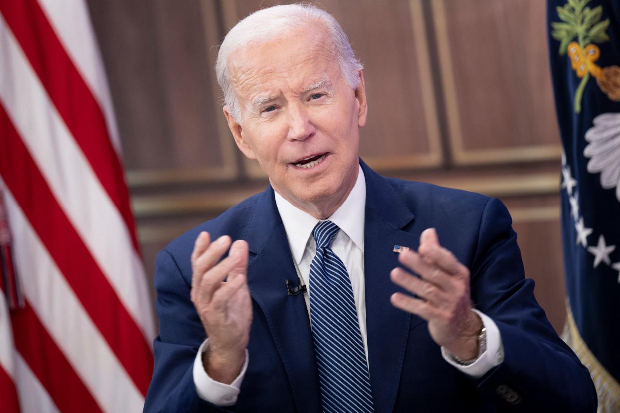 President Biden faces the camera with hands raised to shoulder height, in front of an American flag and a flag with the presidential seal.