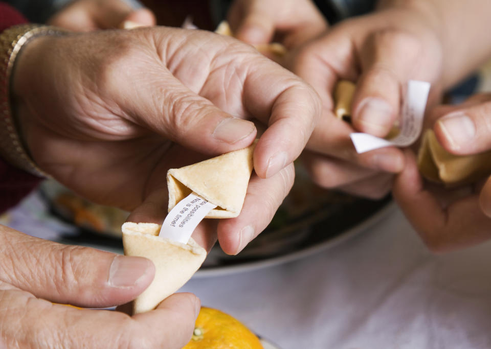 Hands opening fortune cookies with small slips of paper, surrounded by oranges on a table