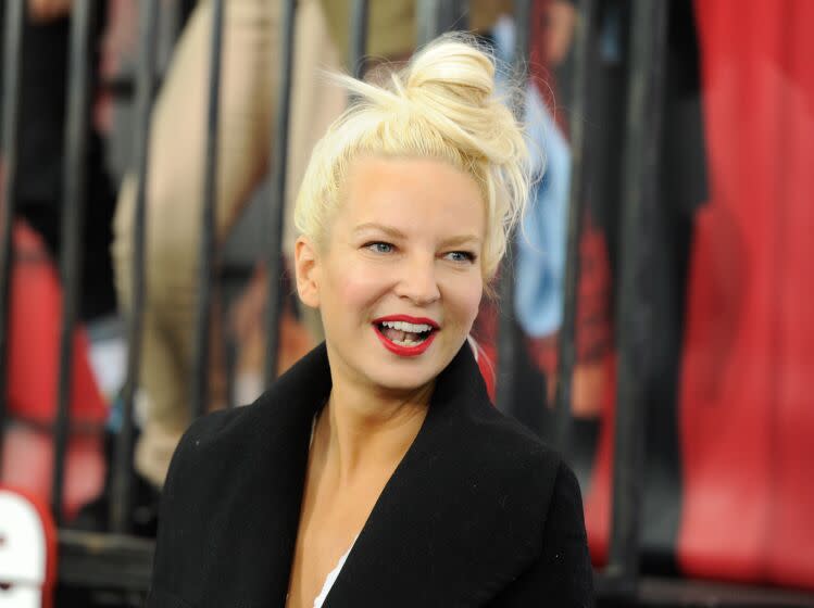 Sia with her hair in a bun wearing red lipstick and a black coat