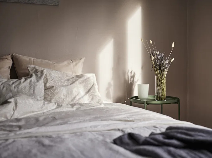 Bedroom scene with unmade bed, side table with a vase of dried flowers and a candle