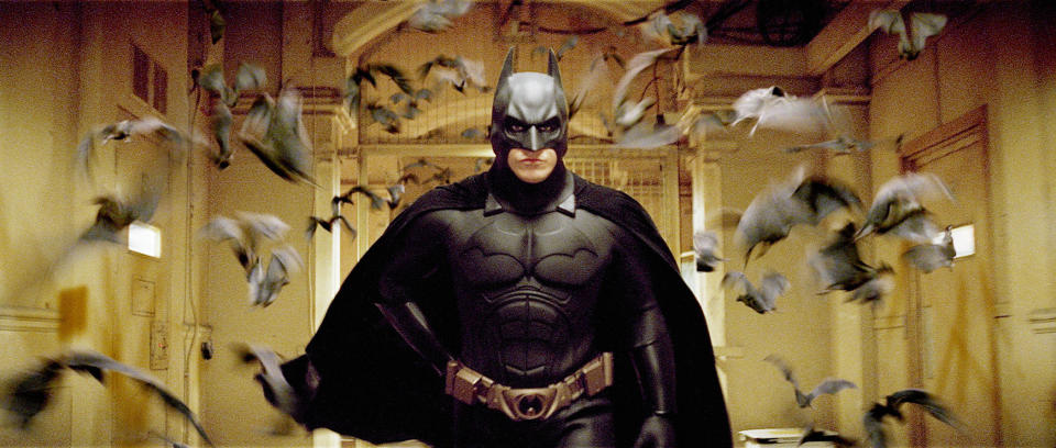 Batman walks through a hallway with bats flying around him, wearing his iconic bat suit in a scene from "Batman Begins"